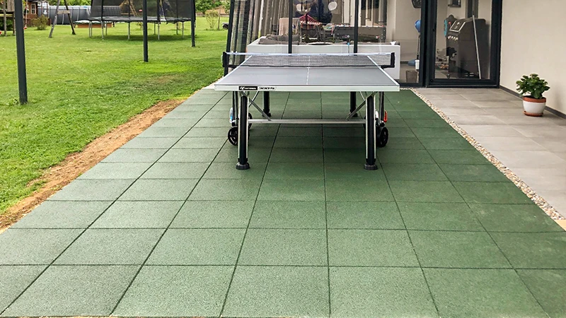 Na terase pokrytej zelenými WARCO dlaždicami z PU viazaného gumového granulátu sa pred luxusným domom nachádza stôl na stolný tenis. V záhrade je trampolína a ďalšie herné prvky, ktoré nabádajú k pohybu a relaxu. Tieto odolné, protišmykové dlaždice poskytujú pohodlie a bezpečnosť. Vďaka pružnému povrchu chránia kĺby, zatiaľ čo ich jednoduchá údržba zaručuje dlhodobo upravený vzhľad.