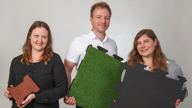 S priateľským úsmevom traja poradcovia spoločnosti WARCO predstavujú podlahové dlaždice z PU viazaného gumového granulátu v hnedej, zelenej a čiernej farbe. Určené na balkóny, poskytujú protišmykový a odolný povrch. Každá verzia má jedinečný dizajn a jednoducho sa kladie. Vďaka odborným znalostiam poradcov si vyberiete ideálne riešenie pre bezpečný a atraktívny balkón. Dlaždice sú nenáročné na údržbu a ponúkajú vysoký komfort.