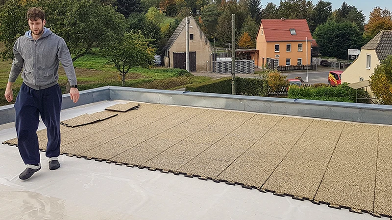 Na fotografii vidno osobu, ktorá ukladá béžové puzzle dosky WARCO na priestranný balkón. Vďaka presnému systému spájania je inštalácia rýchla a jednoduchá. Protišmykový a poveternostne odolný povrch chráni podklad a poskytuje pochôdznu plochu na rôzne využitie. Výsledkom je bezpečný, pohodlný a ľahko udržiavateľný vonkajší priestor, ideálny na oddych. Izolačný účinok navyše pomáha znižovať hluk pri chôdzi.