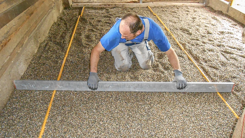 Zhotovenie novej podlahy v konskej stajni: Muž pomocou laty vyrovnáva štrkovú podkladovú vrstvu. Štrk slúži ako lôžko pre montáž boxovej podlahy z gumového granulátu. Plocha je po stranách ohraničená drevenými alebo betónovými stenami. Práca sa vykonáva starostlivo a presne – typické pre odbornú prípravu podlahy vhodnej pre kone.