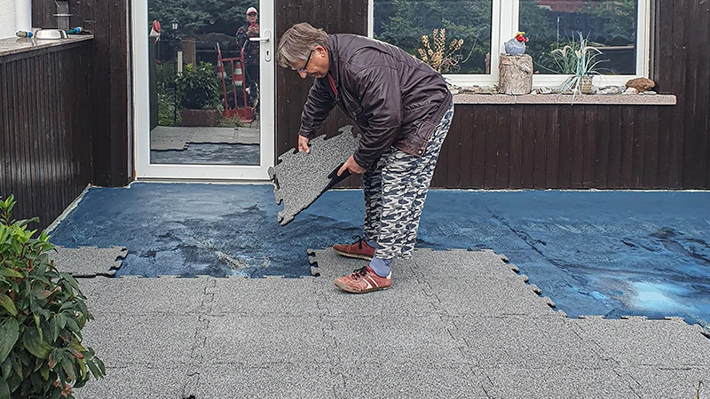 Terasa sa renovuje. Staršia žena pokladá ďalšiu 18 mm hrubú terasovú dlaždicu s puzzle spojom na už čiastočne pokrytú terasu novými dlaždicami. Izolačná fólia slúži ako podklad pre nový podlahový kryt z gumových dlaždíc. Presné spojenie dlaždíc umožňuje jednoduchú a stabilnú pokládku.